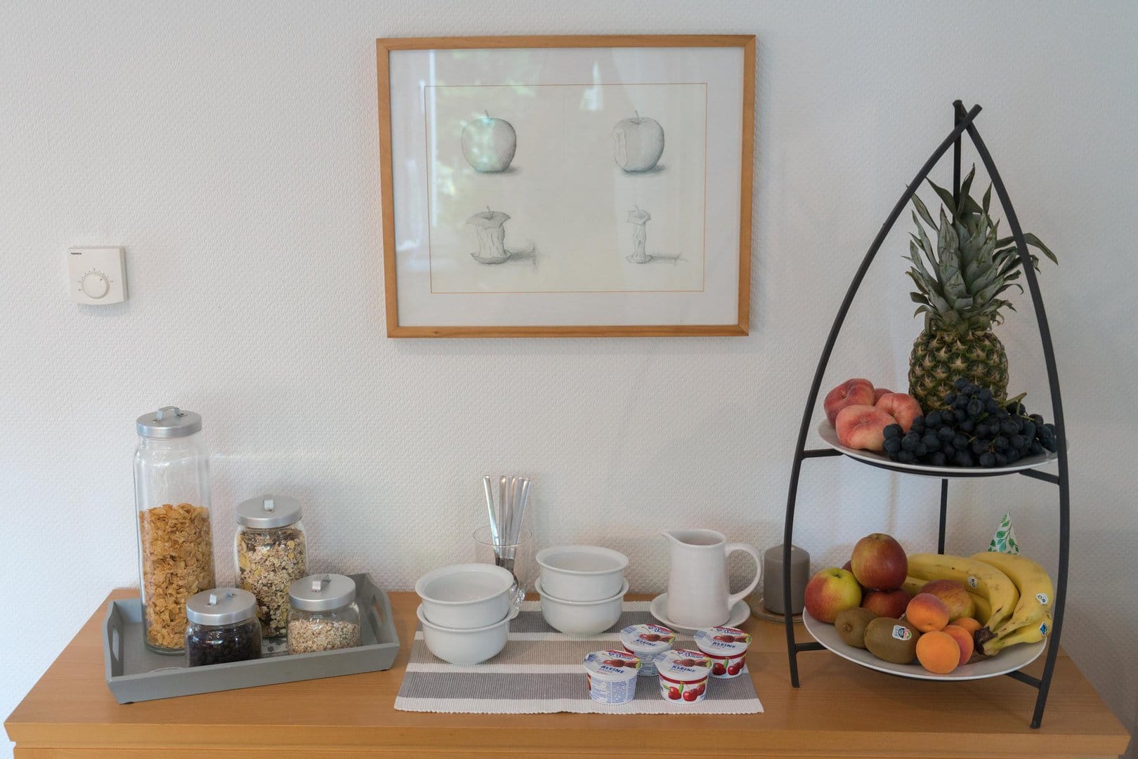 Auf einer Kommode stehen eine Etagere mit diversen Früchten, eine Cerialien-Auswahl, Joghurts, sowie Porzelanschalen und Besteck.