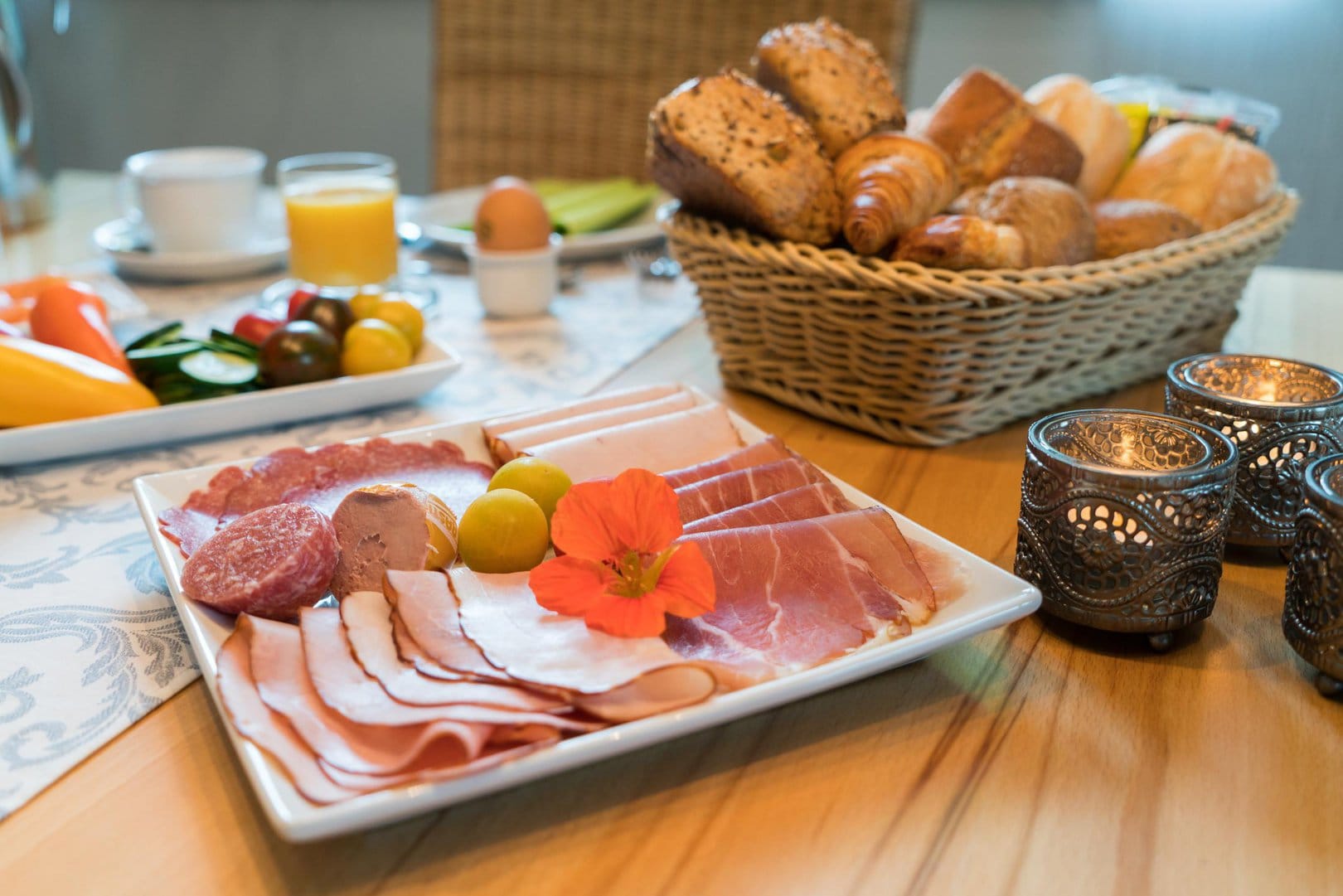 Ein quadratischer Teller mit verschiedenem Wurstaufschnitt. Dahinter steht ein reichhaltiger Brotkorb, sowie ein weiterer Teller mit einer Gemüseauswahl.