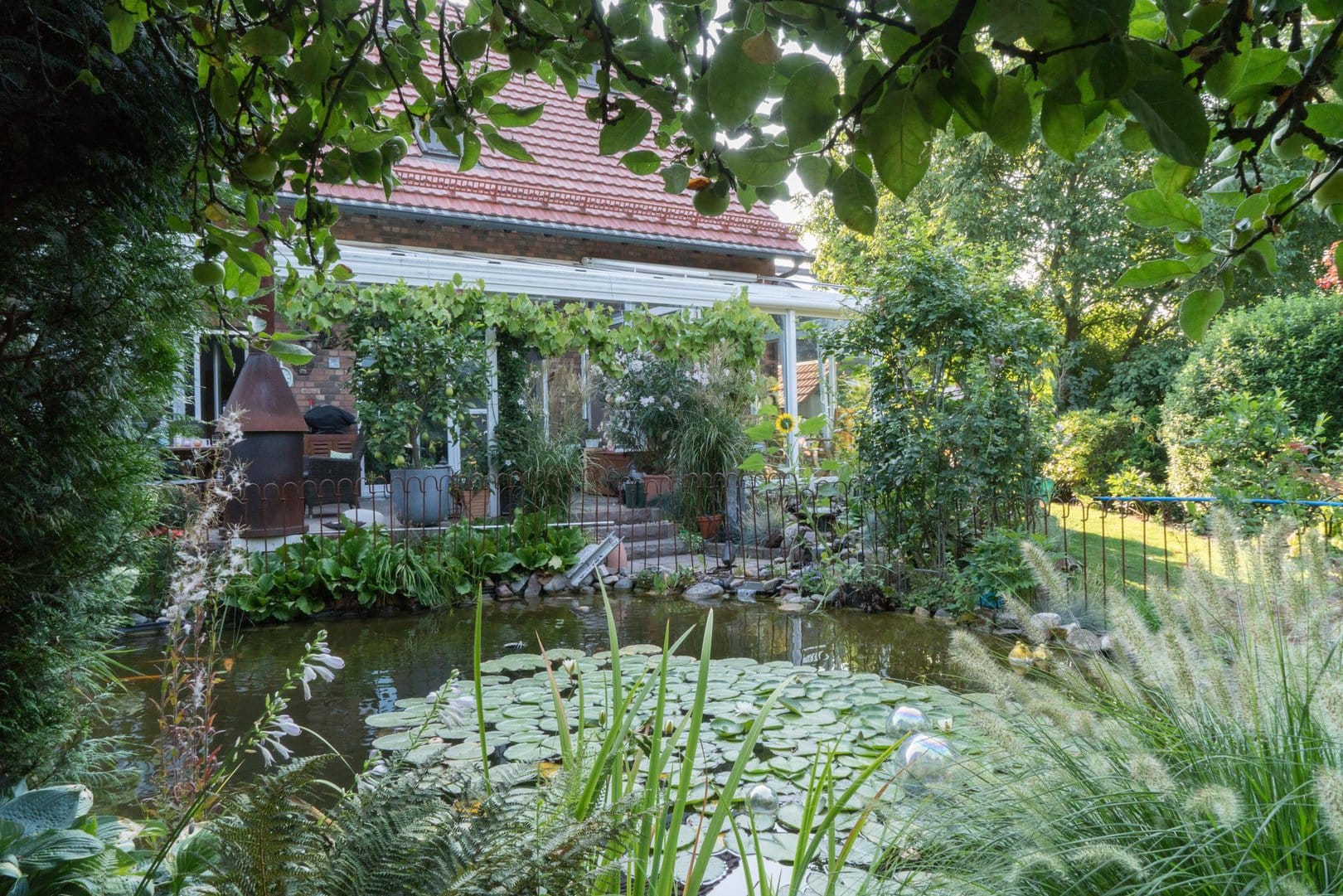 Durch verschiedene Planzen blick man auf den Fischteich, den Rest des Gartens, sowie den Wintergarten.
