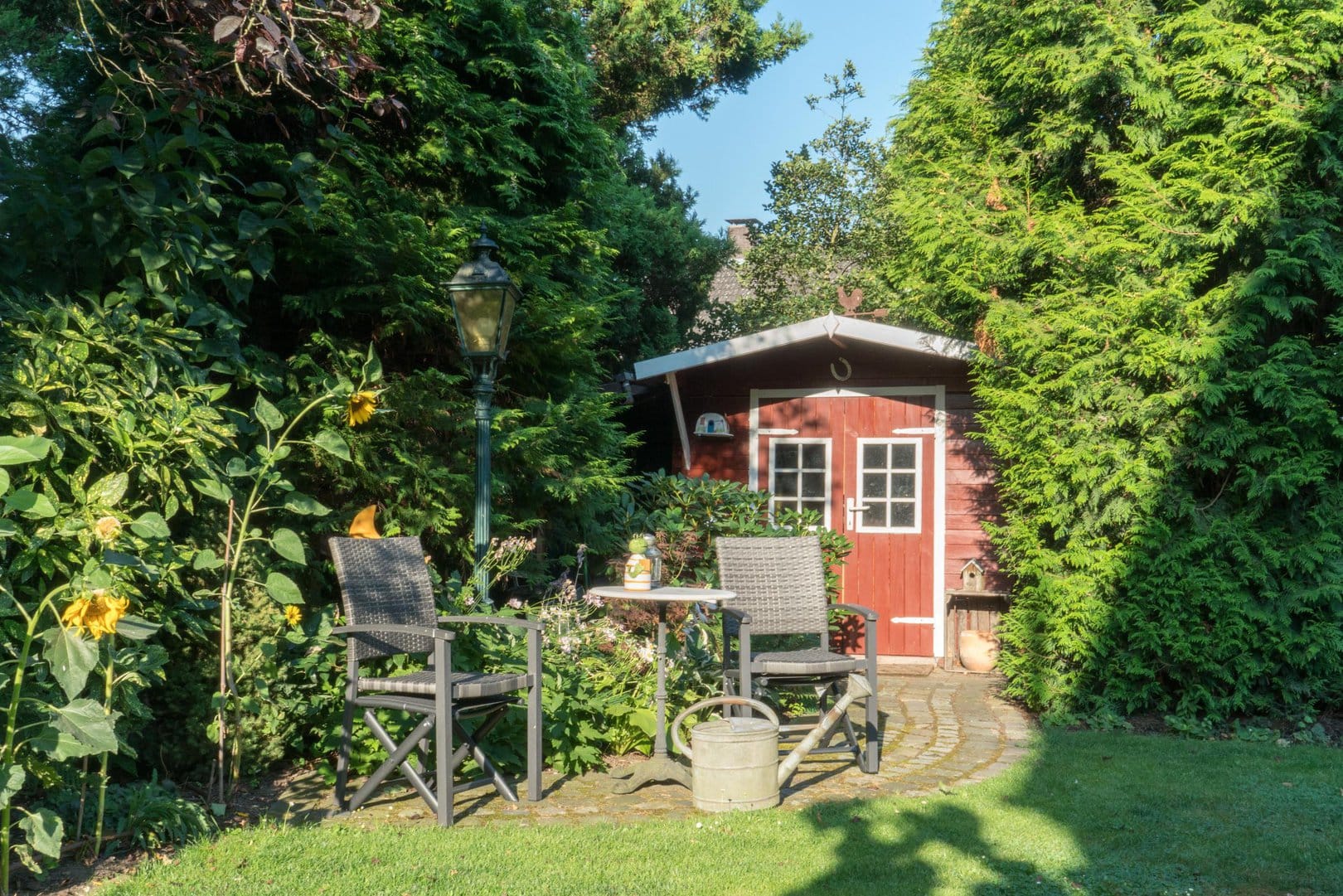 Abwechslungsreich beplanzter Garten mit rotem Gartenhaus und zwei Stühlen und einem kleinen Tisch davor.