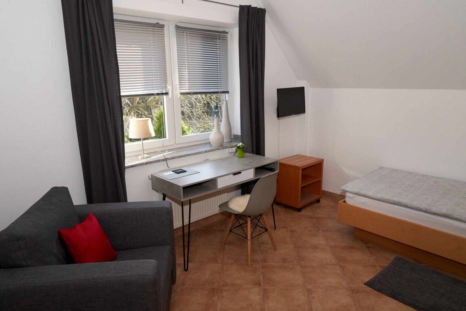 Helles Zimmer mit Einzelbett, Schreibtisch, Stuhl, Sessel, Kleiderschrank und Fernseher. Durch das Fenster sind Bäume zu erkennen.