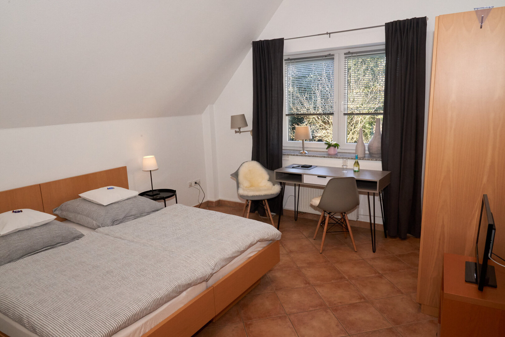 Helles Zimmer mit Doppelbett, Schreibtisch, zwei Stühlen, Kleiderschrank und Fernseher. Durch das Fenster sind Bäume zu erkennen.